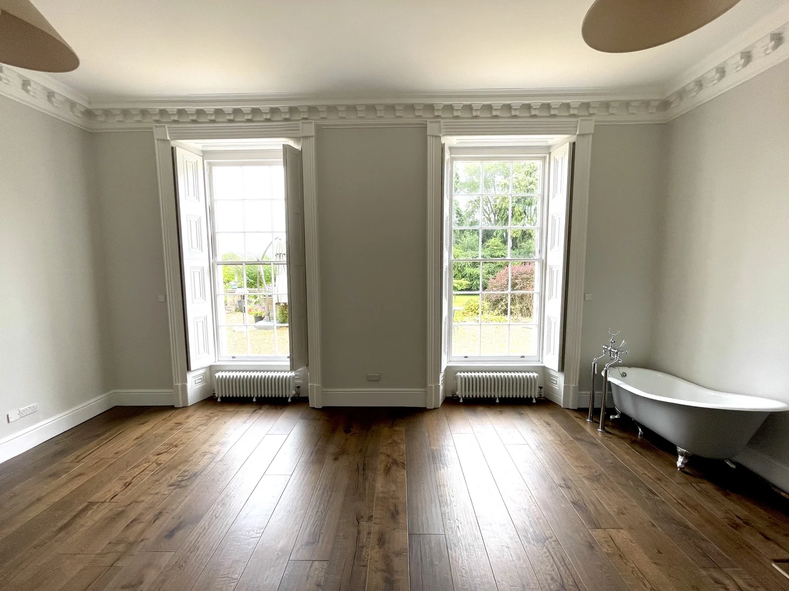 Two tall sash windows above walnut flooring, with a freestanding bath to the right.